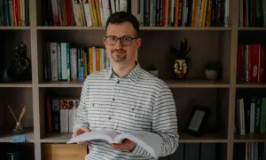 Mężczyzna w okularach, trzymający otwartą książkę, z neutralnym wyrazem twarzy, jakby uczył się, jak nie okazywać emocji na twarzy.